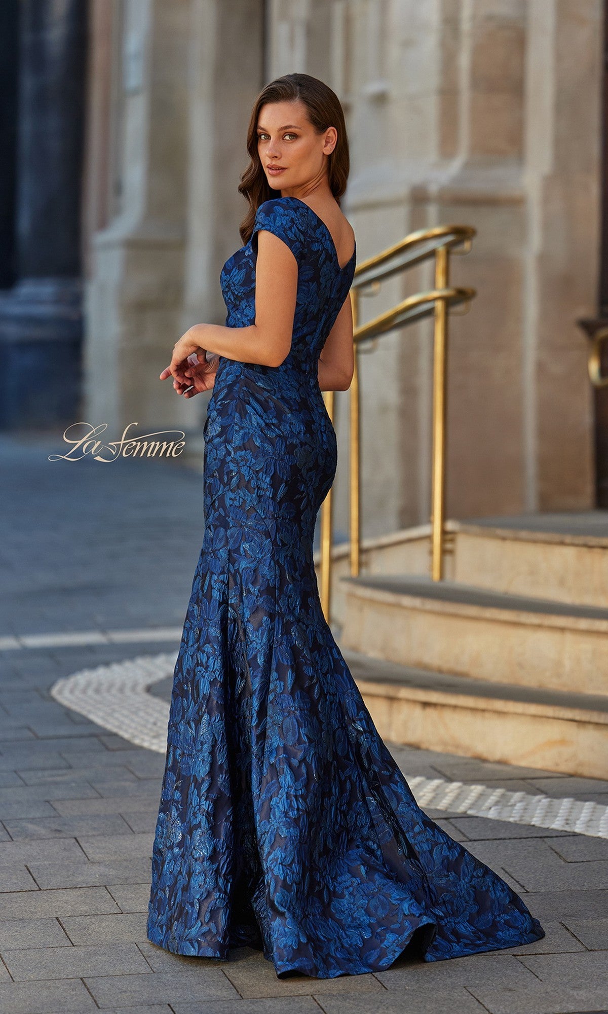 A woman in the La Femme 33578 long formal dress stands on a sidewalk beside a staircase with gold railings.